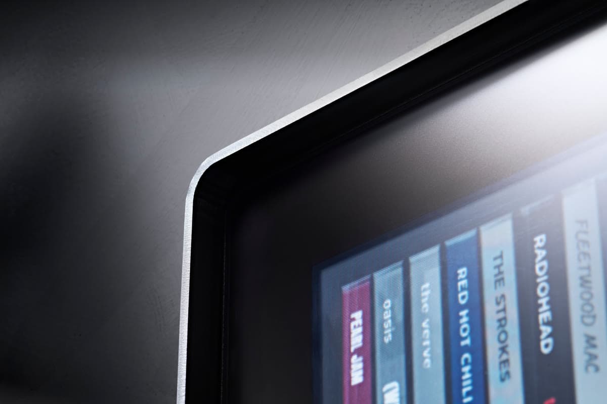 Close-up of Spine's top corner, where the matte glass meets the thin aluminium edge, with album spines in soft focus.